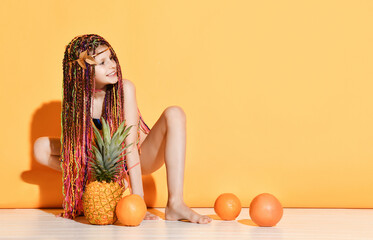 Teenage female with african pigtails, in swimwear. Squatting and smiling, posing with pineapple and...