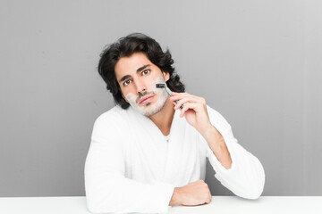 Young caucasian man shaving his beard isolated on a grey background