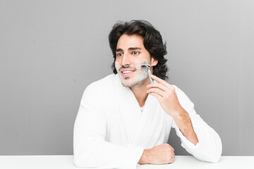 Young caucasian man shaving his beard isolated on a grey background