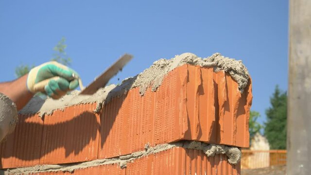 SLOW MOTION, CLOSE UP, DOF: Unrecognizable bricklayer uses a spatula to spread mortar on top of a row of bricks. Detailed shot of builder's hands as he lays bricks on a sunny day. Bricklayer at work.