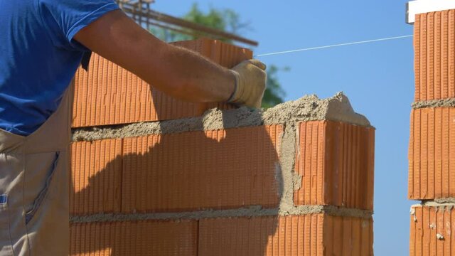 SLOW MOTION, CLOSE UP, DOF: Bricklayer places a brick on top of wet concrete while building a wall at a busy construction site. Unrecognizable worker lays a new brick on top of a wall in progress.
