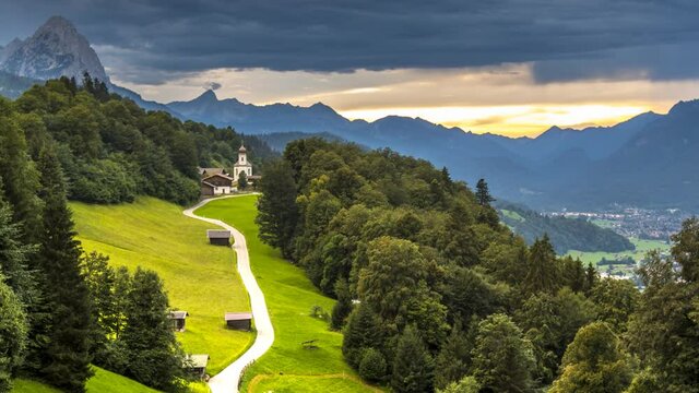Summer time alps mountain nature panoramic landscape near garmisch partenkirchen bavaria germany in backgorun alps mountain in front of church time lapse video in 4k. Sunset meadow.