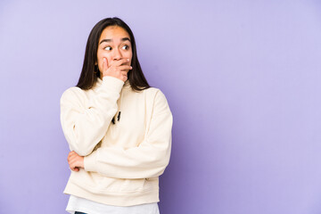 Young woman isolated on a purple background thoughtful looking to a copy space covering mouth with hand.