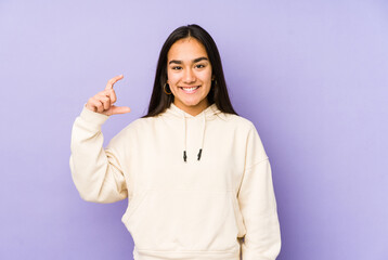 Young woman isolated on a purple background holding something little with forefingers, smiling and confident.