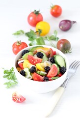 Vegetable berry salad of fresh yellow, red, black tomatoes, cucumbers, onions and blackberries in a bowl on a white background