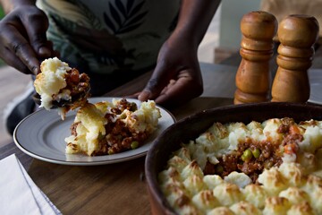 Cottage pie being served up on a plate by a black women.  A wooden salt and pepper shaker sits to the side.  The mince can be seen in the dish.