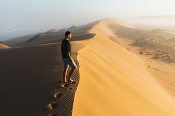 Fototapeta na wymiar Man enjoying sunrise on top of huge sand dune. Beautiful warm sun light. Sahara desert, Morocco.