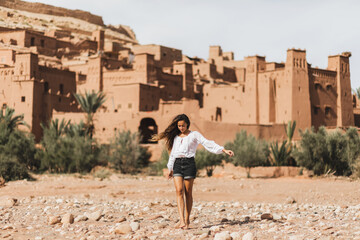 Young curly woman eastern appearance walking on background of kasbah Ait-Ben-Haddou. Travel in Morocco, Ouarzazate. Summer vacations, travel lifestyle concept.
