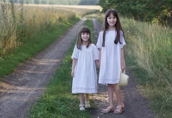 Fotobehang Twee gelukkig meisje op de weg. Schattige lachende meisjes in een witte jurk staan in de buurt van het veld © Albert Ziganshin