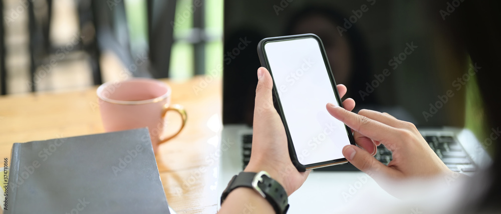 Poster cropped image of a woman's hand is using a mockup smartphone over a computer laptop as a background.