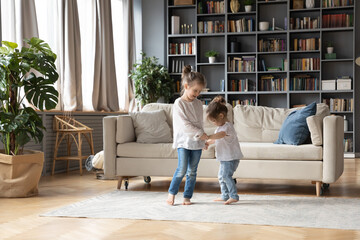 Full length loving small girl holding hands of small toddler sister, dancing barefoot together in floor carpet in living room. Happy two little cute children involved in energetic activity indoors.