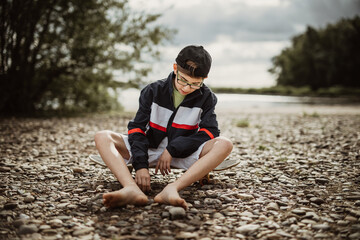 young teen boy child sit on skateboard on stony ground  playing with a stone in his hands and look down enjoy the summer day while exploring the world