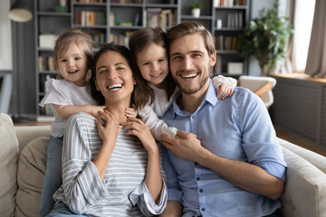 Portrait of affectionate little adorable children girls cuddling happy young parents, relaxing together on comfortable couch in living room, smiling family posing for photo with kids daughters indoors