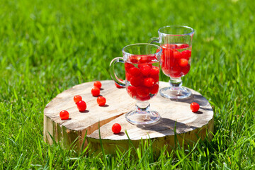 Two glass glasses with cherry berries and water stand on a wooden stand with a crack on the green lawn on a Sunny summer day.