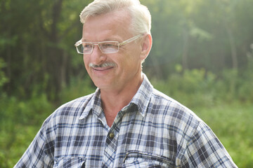 Close-up portrait of handsome gray-haired adult man with mustache and glasses smiling on the background of a summer forest park