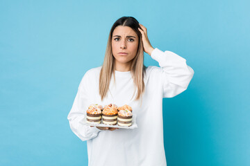 Young caucasian woman holding a sweets cake being shocked, she has remembered important meeting.