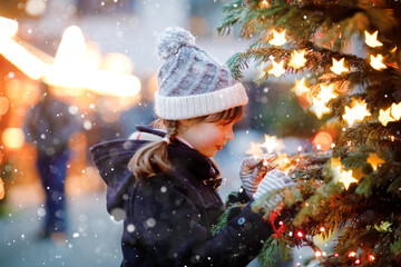 Little cute kid girl having fun on traditional Christmas market during strong snowfall. Happy child enjoying traditional family market in Germany. Schoolgirl standing by illuminated xmas tree.