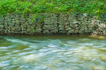 A fast-flowing river enclosed by a steep bank with wire protection from shattering. Reinforced bank with stones against soil slipping into the river and erosion.