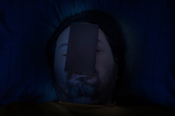 A middle-aged bearded man sleeps on a pillow late at night with a mobile phone lying on his face....