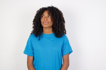 Fototapeta premium Cheerful positive Young african woman with curly hair wearing casual blue shirt over white background, looking aside into empty space thoughtful
