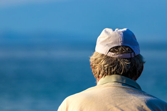 Close Up Shot Of The Back Of A Senior Man's Head With Gray Hair Wearing A Cup Looking The Sea.