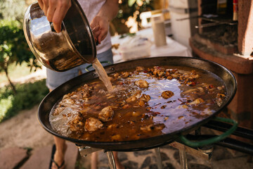 Young woman pouring seafood broth into the paella pan to prepare a typical Spanish paella
