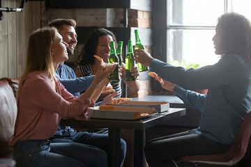 Overjoyed diverse people clinking with beer bottles, eating pizza, happy excited young men and women enjoying weekend in cafe or pizzeria, having fun together, celebrating event in bar