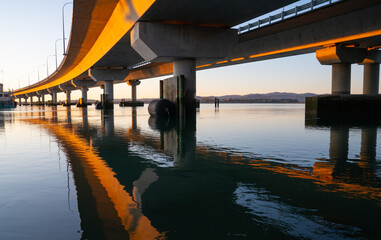 Morning sun strikes side Tauranga Harbour Bridge in golden hue reflected leading lines into calm water below