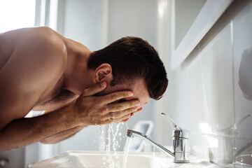 Handsome young naked man is washing his face in the bathroom at home.