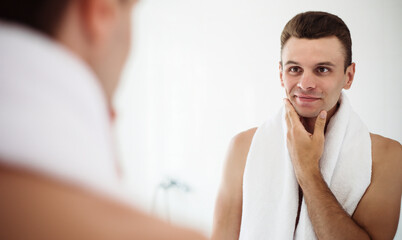 Fototapeta premium Handsome young man shaving his beard in the bathroom. Portrait of a stylish naked bearded man examining his face in-home mirror.