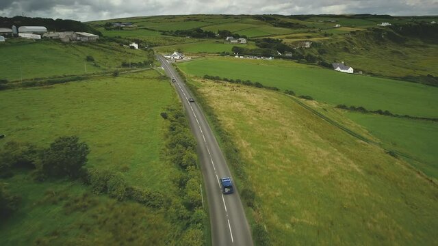 Ireland rural road aerial shot: cars riding near crossway. Cottages, meadows, fields on hills on cloudy summer day. Irish beautiful landscape of countryside. Footage view in FullHD
