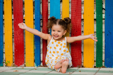 Joyful little girl with open hands laughs and sends love to everyone. Behind her is a multi-colored...