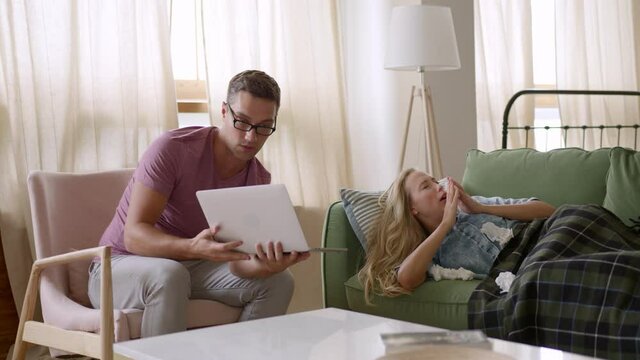 Young Married Couple Have Video Telemedicine Call Using Web Camera On Laptop At Home. Blonde Woman Lays On Sofa Under The Blanket, Sneezing, Feeling Sick, Man Holds Laptop And Talks With Doctor. 