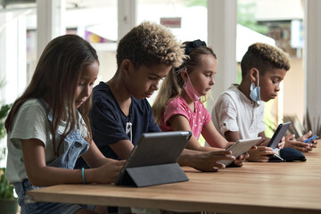 Fototapeta premium Four diverse kids wear facemasks sit at table use wireless gadgets ignoring each other prefer internet games and virtual communication. Alpha generation and modern technology overuse, phubbing concept