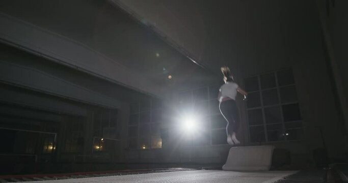 Girl gymnast performs the right jumping somersault in the air on a trampoline in sportswear in a dark gym with single bright spotlight. Night gymnastics training of a girl in a sports hall at night.
