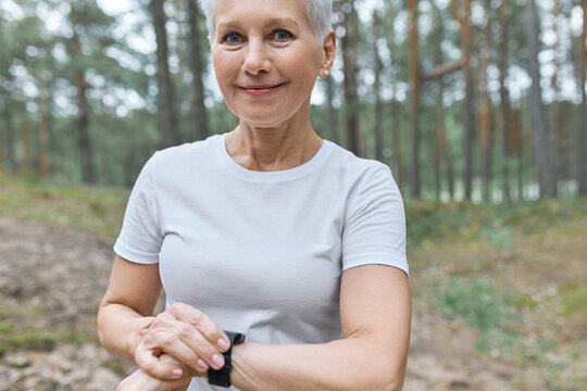 Cropped Shot Of Beautiful Middle Aged Woman In White T-shirt Adjusting Smart Watch On Her Wrist, Tracking Heart Rate, Counting Calories And Steps During Workout, Happy With Progress, Smiling At Camera