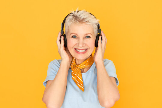 Joyful happy mature short haired woman smiling broadly posing against yellow background in wireless headphones, enjoying clean high quality sound, listening to music online, having cheerful look