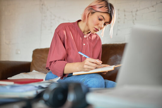 Concentrated young woman copywriter with pink hair working from home making notes in copybook. Cute student girl putting down ideas, writing essay, having focused facial expression, sitting on couch