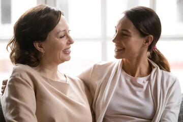 Head shot smiling beautiful brown-haired mature woman communicating with happy grown daughter, sitting together on couch indoors. Cheerful female family sharing news, enjoying pleasant conversation.