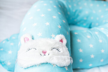 Light blue supporting pillow with stars on white sheet in bed for comfortable sleep of pregnant woman. Funny sleeping mask in form of bunny,cat with eyes,ears,mouth. Accessories for expectant mothers