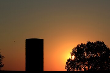 Sunset over a farm out in the country with a Silo, Grain Bin, and a tree with silhouette's.