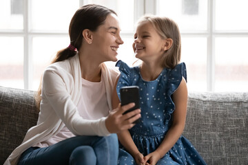 Fototapeta premium Happy young mixed race woman recording funny video on smartphone with little cute sister or daughter. Laughing preschool small child watching media content on cellphone with caring mum or nanny.