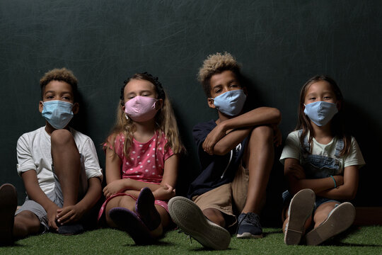 Four Multinational Children Wears Surgical Protective Cloth Facemasks School Age Girls And Boys Look At Camera Posing Sitting Leaned At Dark Grey Wall Background. Corona Virus And Kids Concept