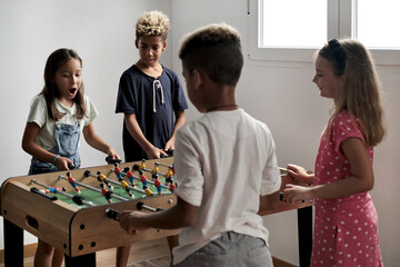Group of multi cultural cute children stand indoor spend playtime with friends playing foosball together. Tabletop version of soccer lovers. Having fun and recreation of alpha generation kids concept