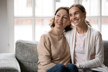 Dreamy bonding different female generations mixed race family sitting on couch, looking in...
