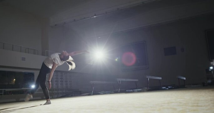The girl gymnast performs the coup exercise on one arm with the help of force in the legs in a dark gym with single bright spotlight. Night gymnastics training of a girl in a sports hall at night.