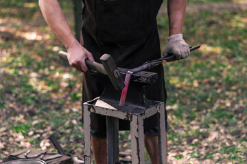 Obraz premium a young male blacksmith forges a product in nature, a lit hearth and a small portable hand anvil. Hammer work, metallurgy. Handmade metal. Blacksmiths Festival of craftsmanship, street exhibition.