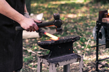 a young male blacksmith forges a product in nature, a lit hearth and a small portable hand anvil....