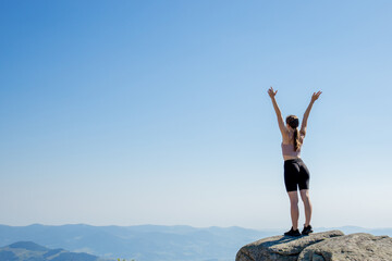 Obraz premium The young girl at the top of the mountain raised her hands up on blue sky background. The woman climbed to the top and enjoyed her success. Back view