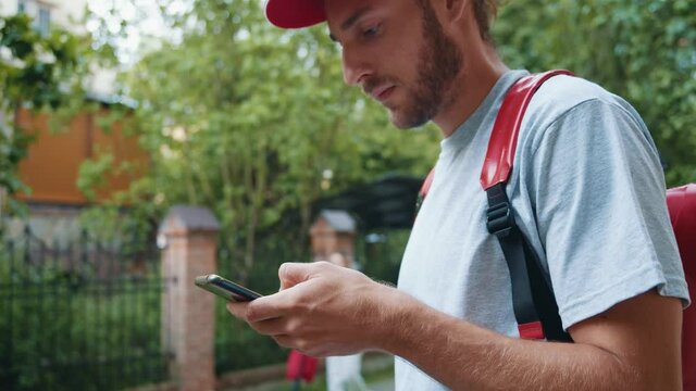 Urban courier delivery services worker in uniform with backpack searching customer's adress on mobile phone device outdoor. Summer day. Food delivery.